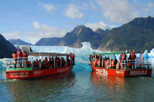 Two red excursion boats approach a glacier amid floating icebergs on a bright day in Chilean Patagonia. 