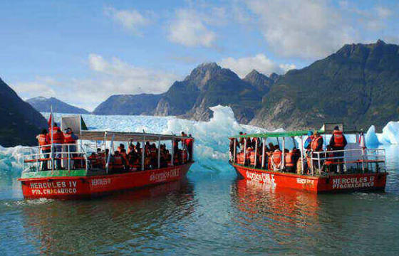 Two red excursion boats approach a glacier amid floating icebergs on a bright day in Chilean Patagonia. 
