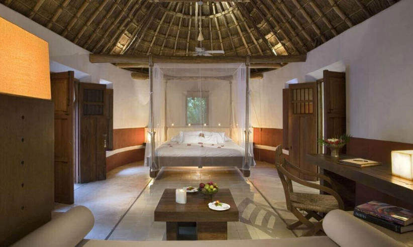 Spacious interior of a luxury Mayan-style villa with palapa roof, canopy bed, and natural wood furnishings in Mexico. 