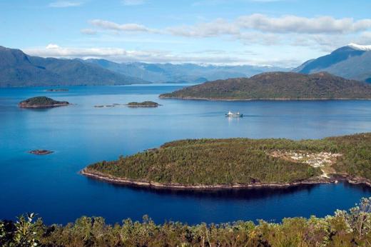 A cruise ship navigates the deep blue waters of Chile's fjords, weaving between lush green islands and distant Andean peaks. 