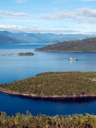 A cruise ship navigates the deep blue waters of Chile's fjords, weaving between lush green islands and distant Andean peaks. 
