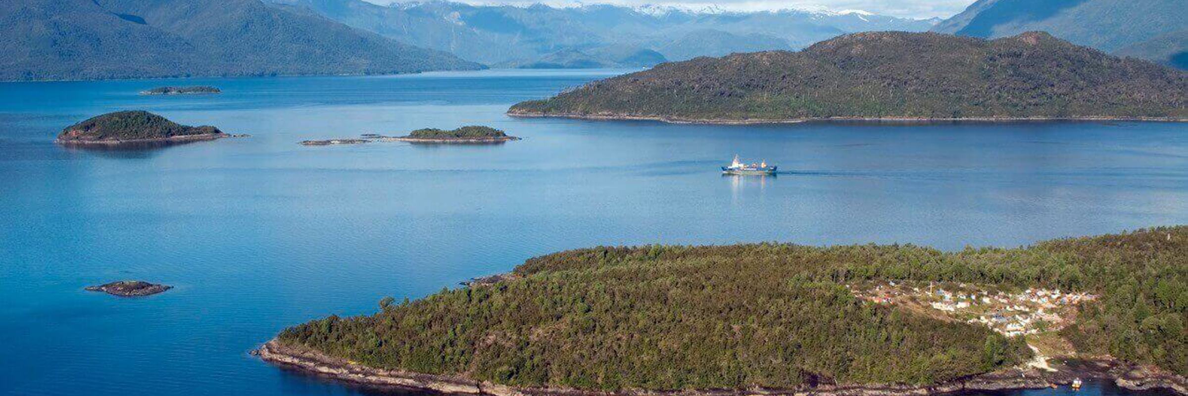 A cruise ship navigates the deep blue waters of Chile's fjords, weaving between lush green islands and distant Andean peaks. 