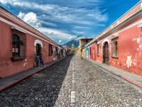 Cobblestone street in Antigua, Guatemala with pastel houses and a distant volcano under blue skies. 