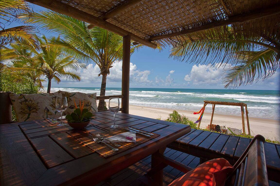 Table with ocean view at beachfront restaurant in Bahia, Brazil, shaded by palm trees and thatch roof. 
