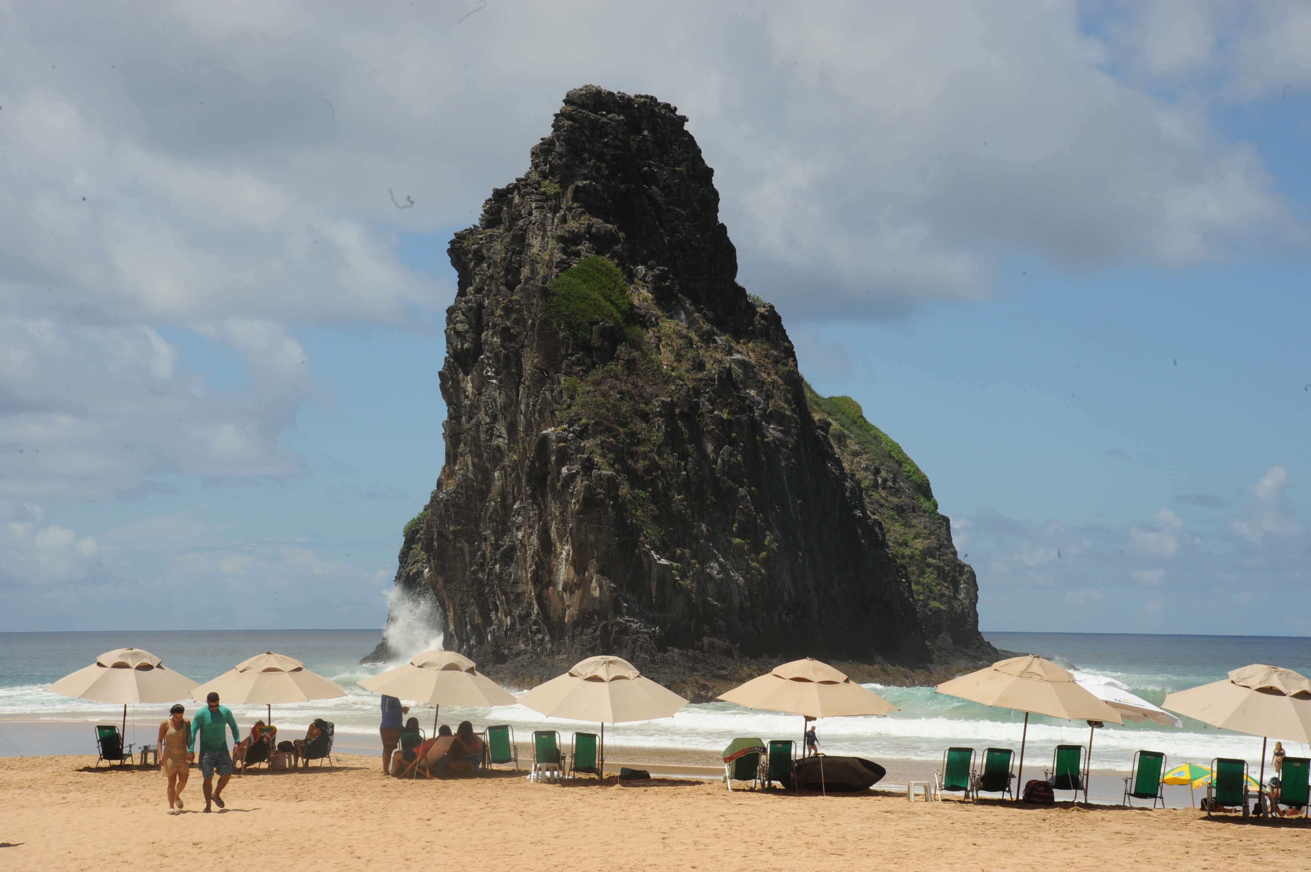 Dramatic beach rock formation at Praia do Cachorro in Fernando de Noronha, with parasols and people relaxing in the sand. 
