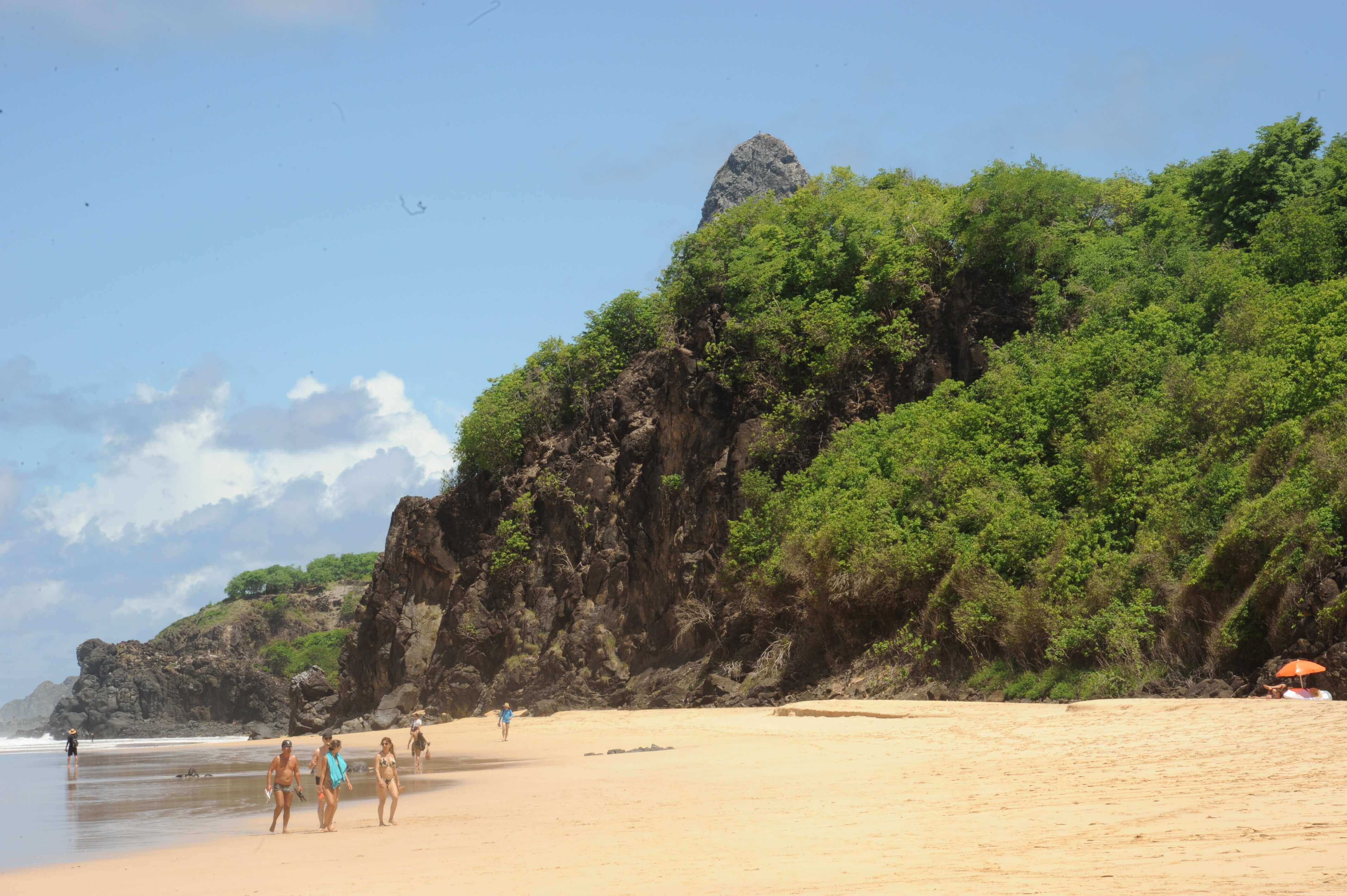 Lush tropical vegetation meets golden sand at Praia do Meio in Fernando de Noronha, with visitors walking along the beach. 