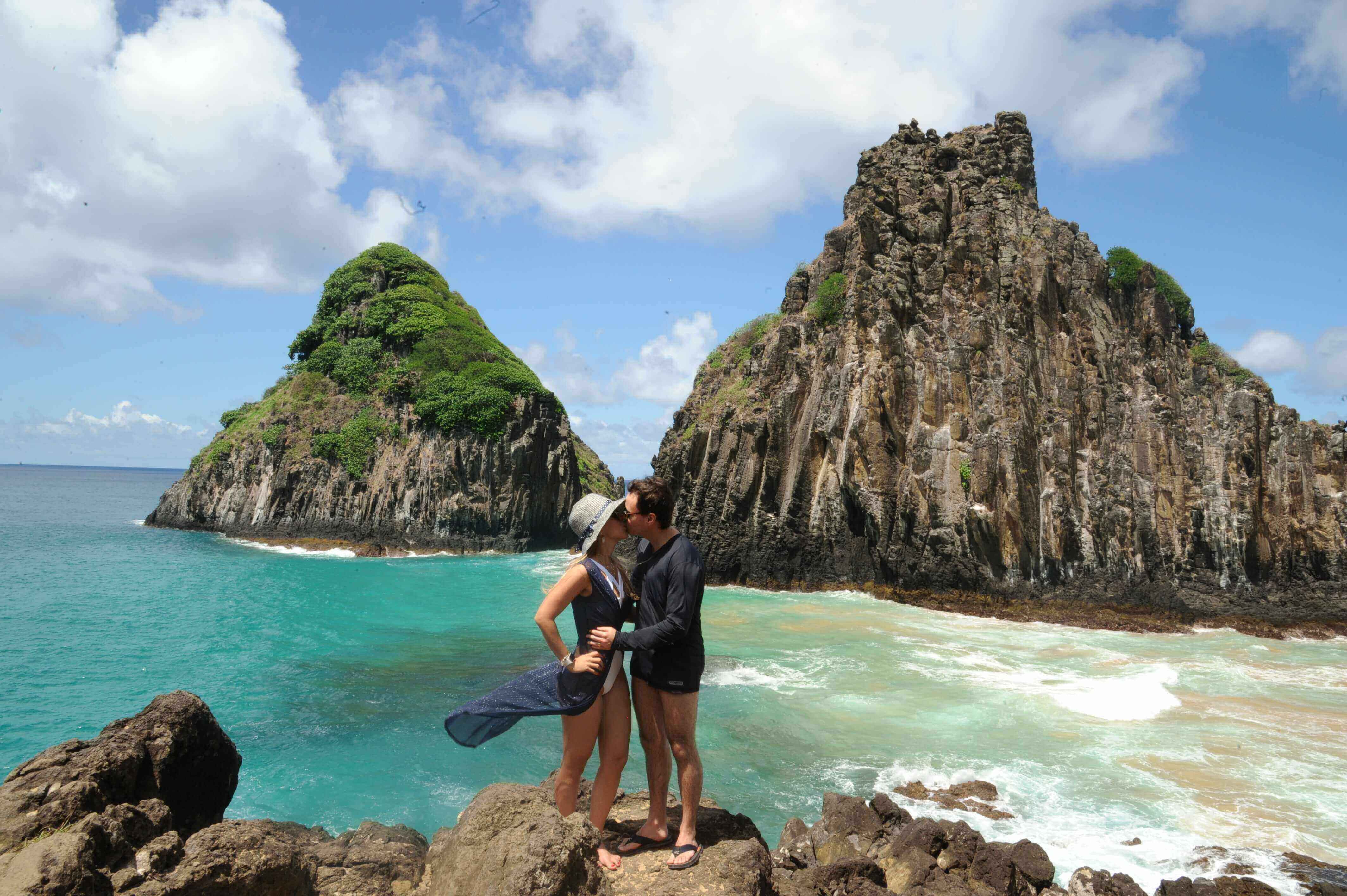 Couple standing on rocks with view of Baia dos Porcos and iconic twin peaks at Fernando de Noronha, Brazil. 
