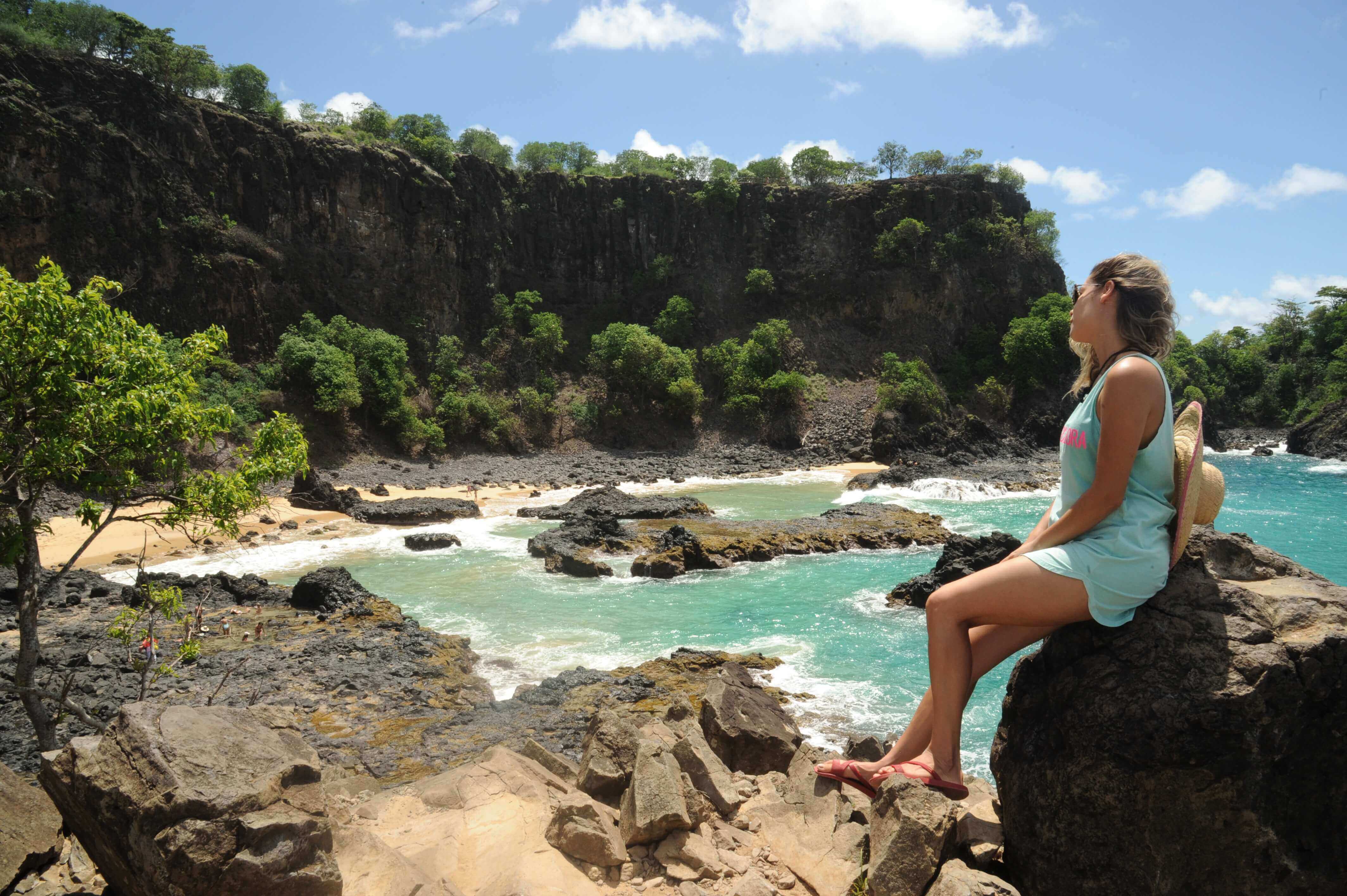 Girl seated on a cliff overlooking turquoise waters and rugged coastline in Fernando de Noronha. 