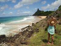 Woman with straw hat overlooking rocky coastline and Morro do Pico at Fernando de Noronha, Brazil. 