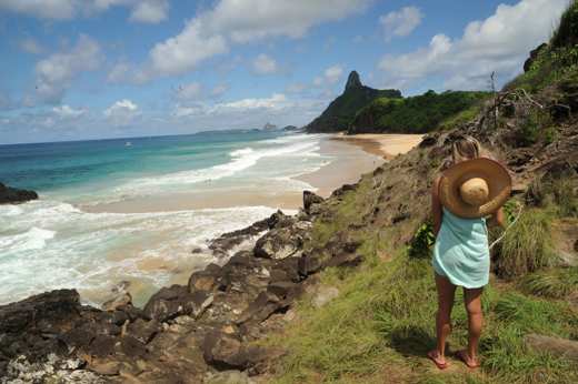 Woman with straw hat overlooking rocky coastline and Morro do Pico at Fernando de Noronha, Brazil. 