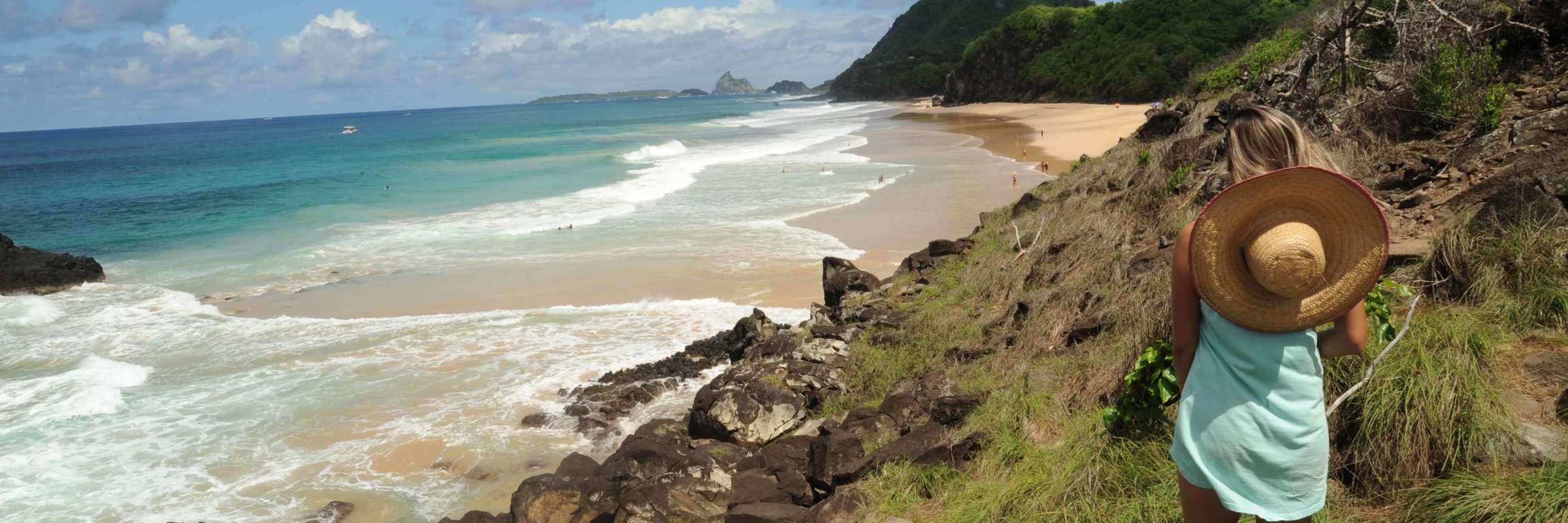 Woman with straw hat overlooking rocky coastline and Morro do Pico at Fernando de Noronha, Brazil. 