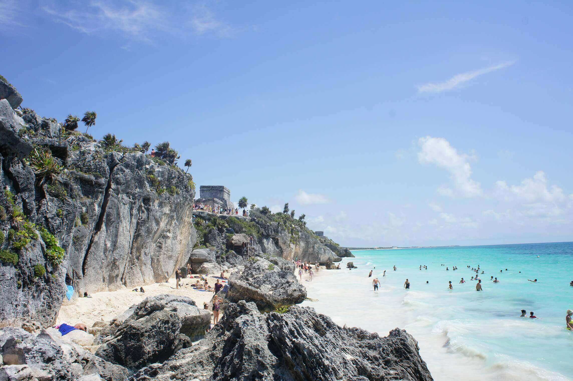 Visitors enjoy Tulum's beach with turquoise waters, white sand, and ancient ruins perched on the cliffside above. 