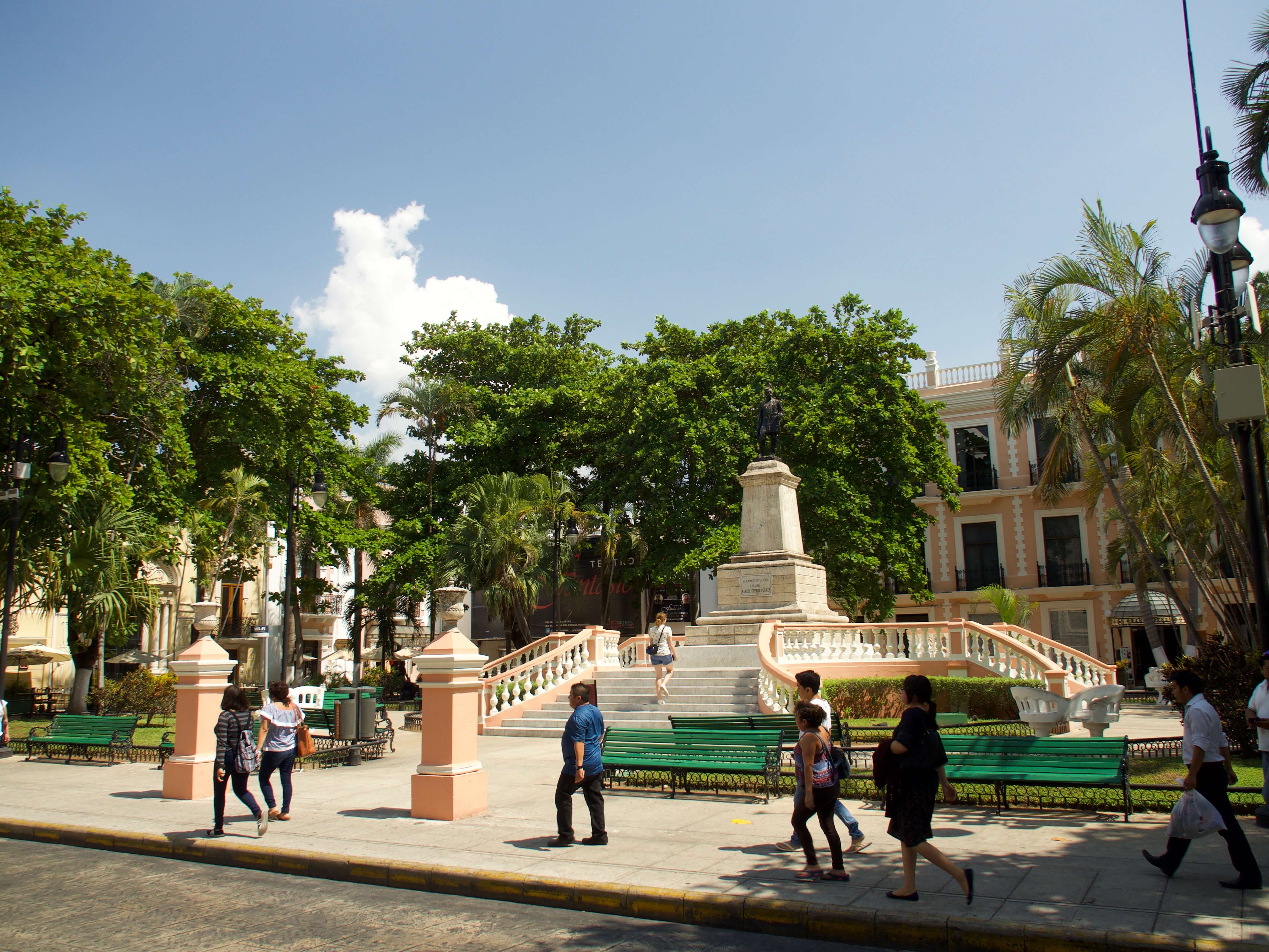 View of the vibrant Plaza Grande (Zocalo) in Merida, Yucatan, Mexico, with locals strolling past green benches and colonial buildings. 