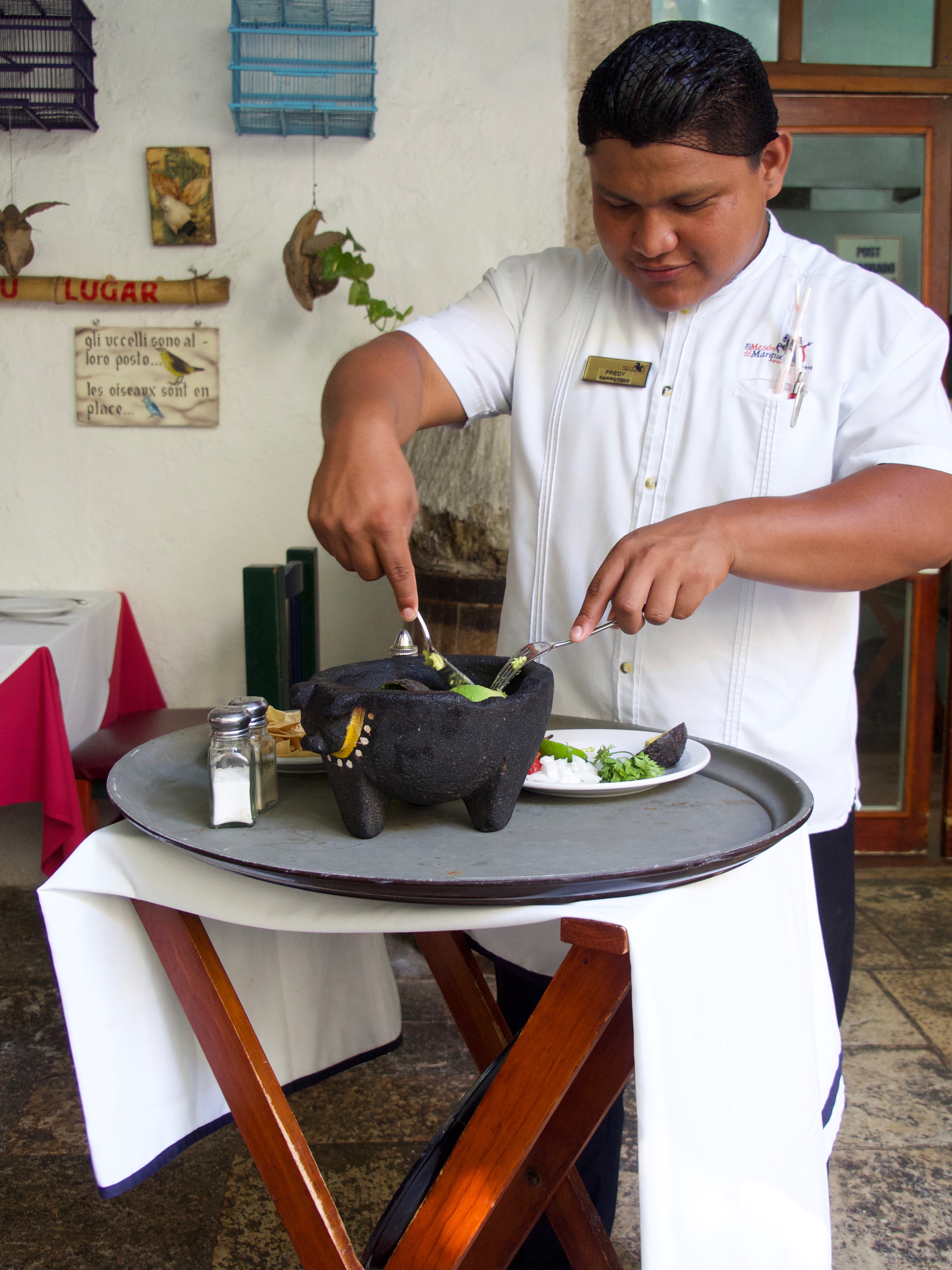 Server prepares fresh guacamole tableside using a traditional molcajete at a Mexican restaurant. 