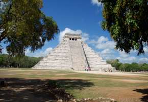 The Temple of Kukulkan, also known as El Castillo, stands tall at Chichen Itza under a bright blue Yucatan sky surrounded by trees. 