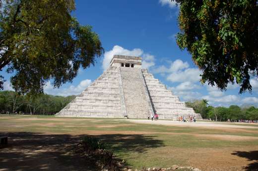 The Temple of Kukulkan, also known as El Castillo, stands tall at Chichen Itza under a bright blue Yucatan sky surrounded by trees. 