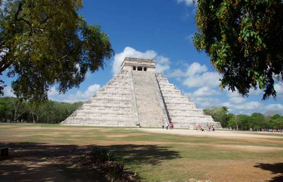 The Temple of Kukulkan, also known as El Castillo, stands tall at Chichen Itza under a bright blue Yucatan sky surrounded by trees. 