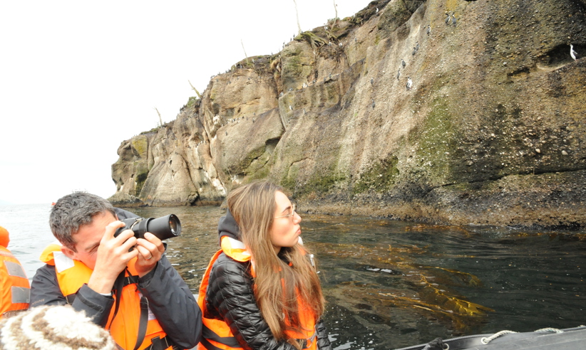 Two cruise passengers in a Zodiac take photos of dramatic coastal cliffs during a  Patagonia expedition in Chile. 