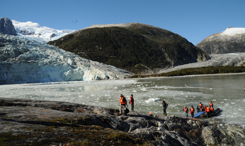 Cruise passengers in life vests explore the rocky glacier shoreline in Chilean Patagonia under clear blue skies. 