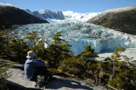 A lone hiker rests on a rock ledge, gazing at a massive glacier framed by snowy peaks in Chilean Patagonia. 