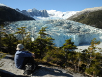 A lone hiker rests on a rock ledge, gazing at a massive glacier framed by snowy peaks in Chilean Patagonia. 