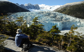 A lone hiker rests on a rock ledge, gazing at a massive glacier framed by snowy peaks in Chilean Patagonia. 