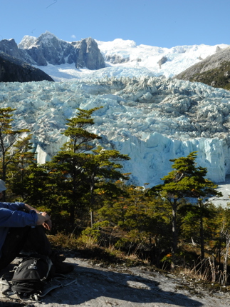 A lone hiker rests on a rock ledge, gazing at a massive glacier framed by snowy peaks in Chilean Patagonia. 