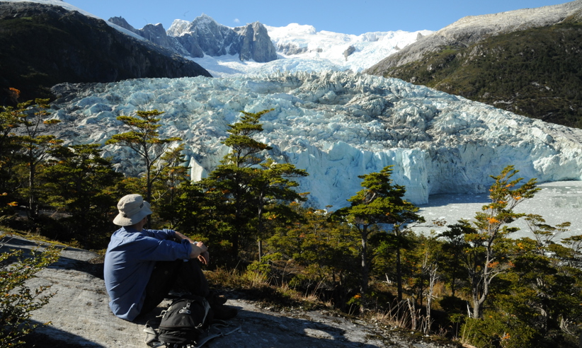 A lone hiker rests on a rock ledge, gazing at a massive glacier framed by snowy peaks in Chilean Patagonia. 