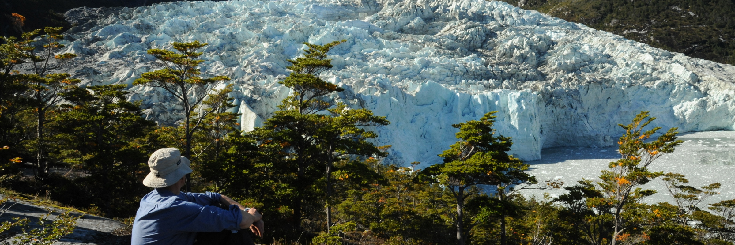A lone hiker rests on a rock ledge, gazing at a massive glacier framed by snowy peaks in Chilean Patagonia. 