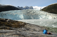 A tourist sits on rocky ground gazing at a towering glacier and jagged snowy peaks in remote Patagonia, Chile. 