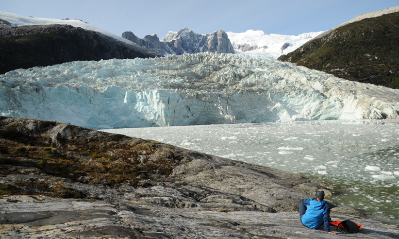 A tourist sits on rocky ground gazing at a towering glacier and jagged snowy peaks in remote Patagonia, Chile. 