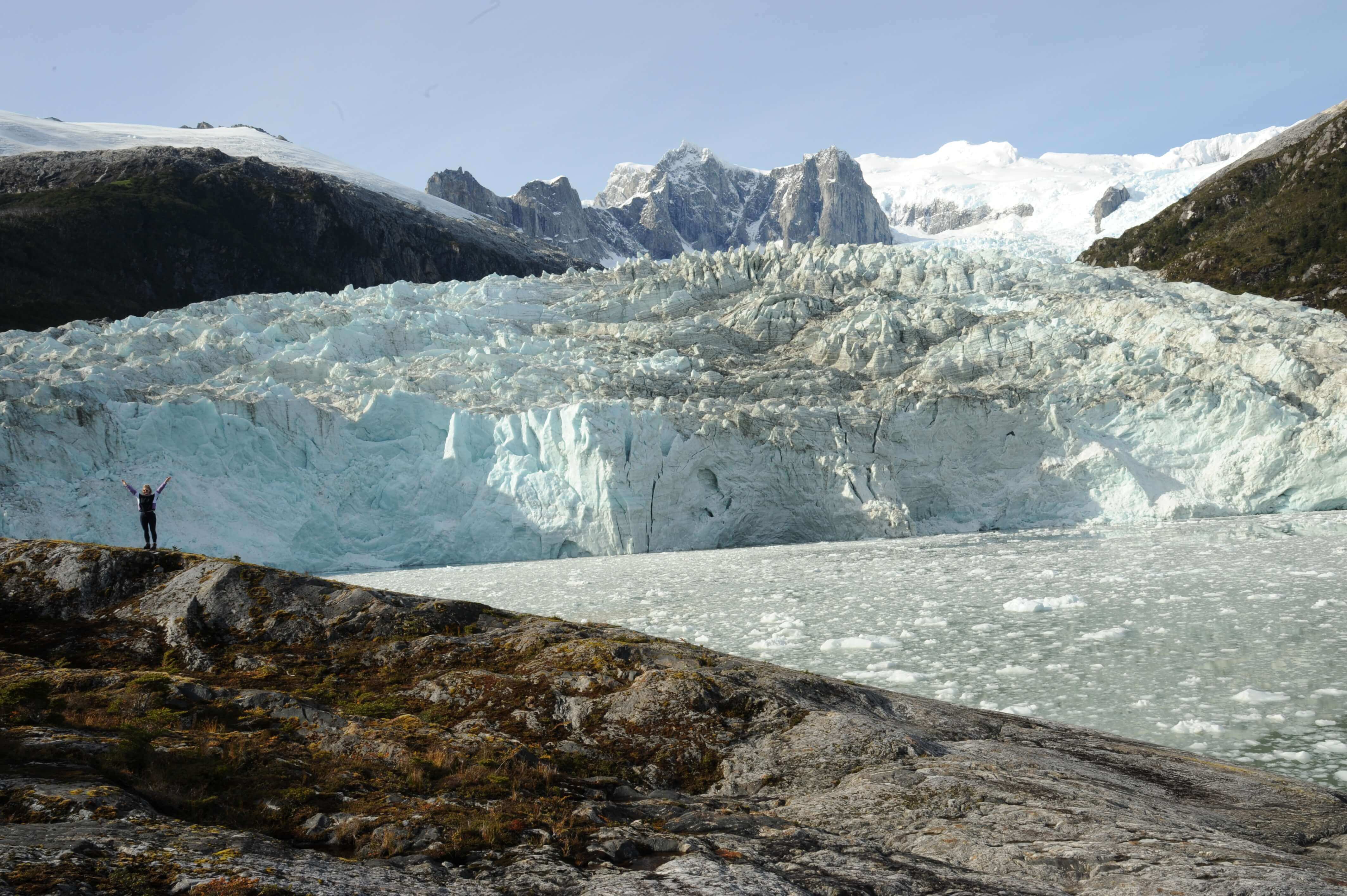 A hiker approaches the massive glacier wall in Tierra del Fuego, framed by rugged peaks and brilliant blue ice. 