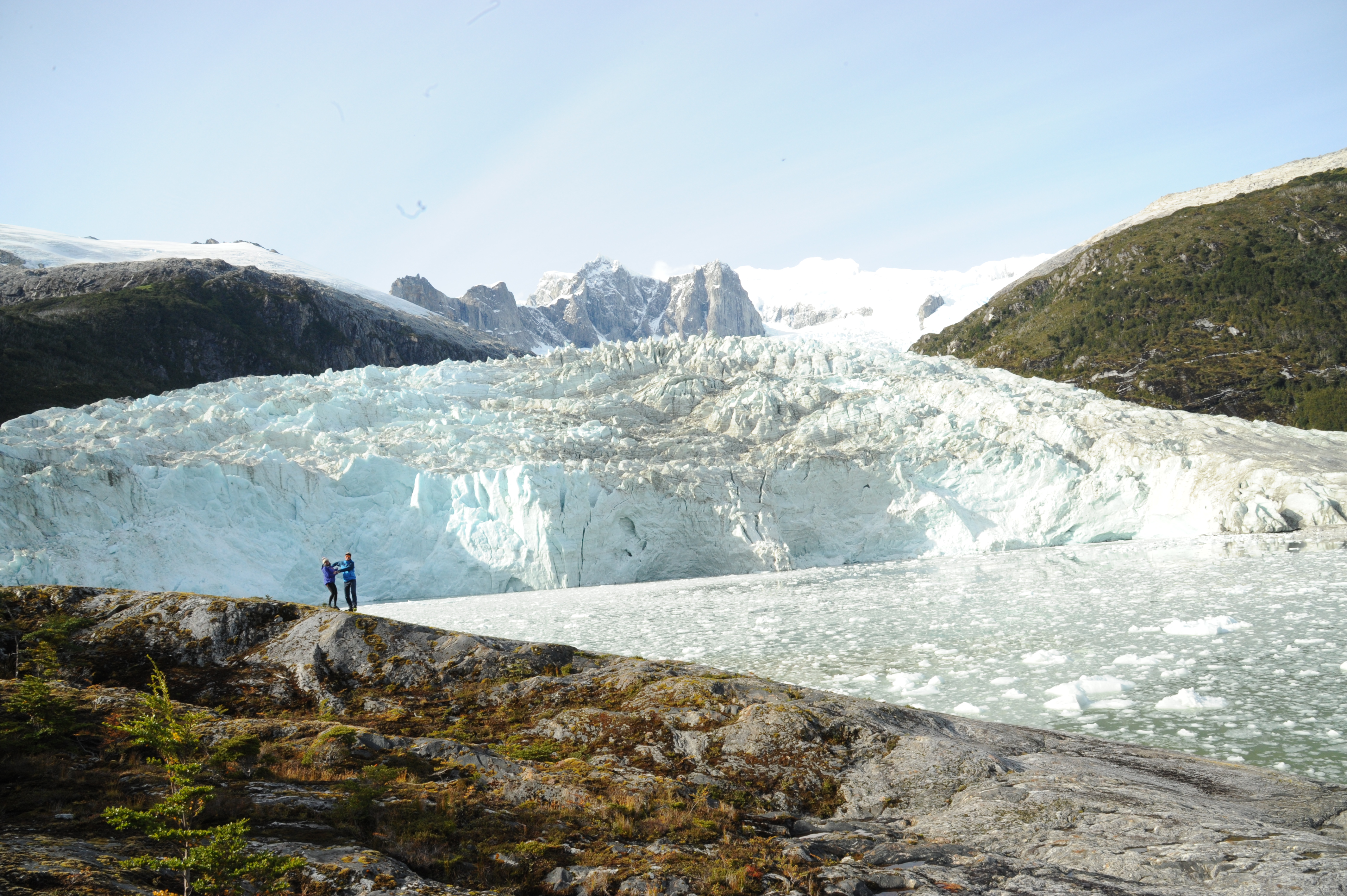 Two travelers stand on rokcy terrain facing a towering blue glacier beneath jagged peaks in remote Patagonia, Chile. 