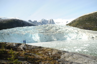 Two travelers stand on rokcy terrain facing a towering blue glacier beneath jagged peaks in remote Patagonia, Chile. 