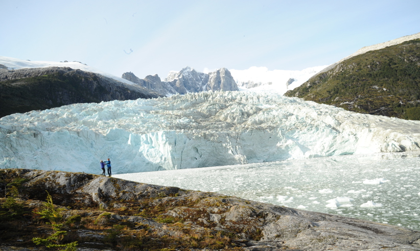 Two travelers stand on rokcy terrain facing a towering blue glacier beneath jagged peaks in remote Patagonia, Chile. 