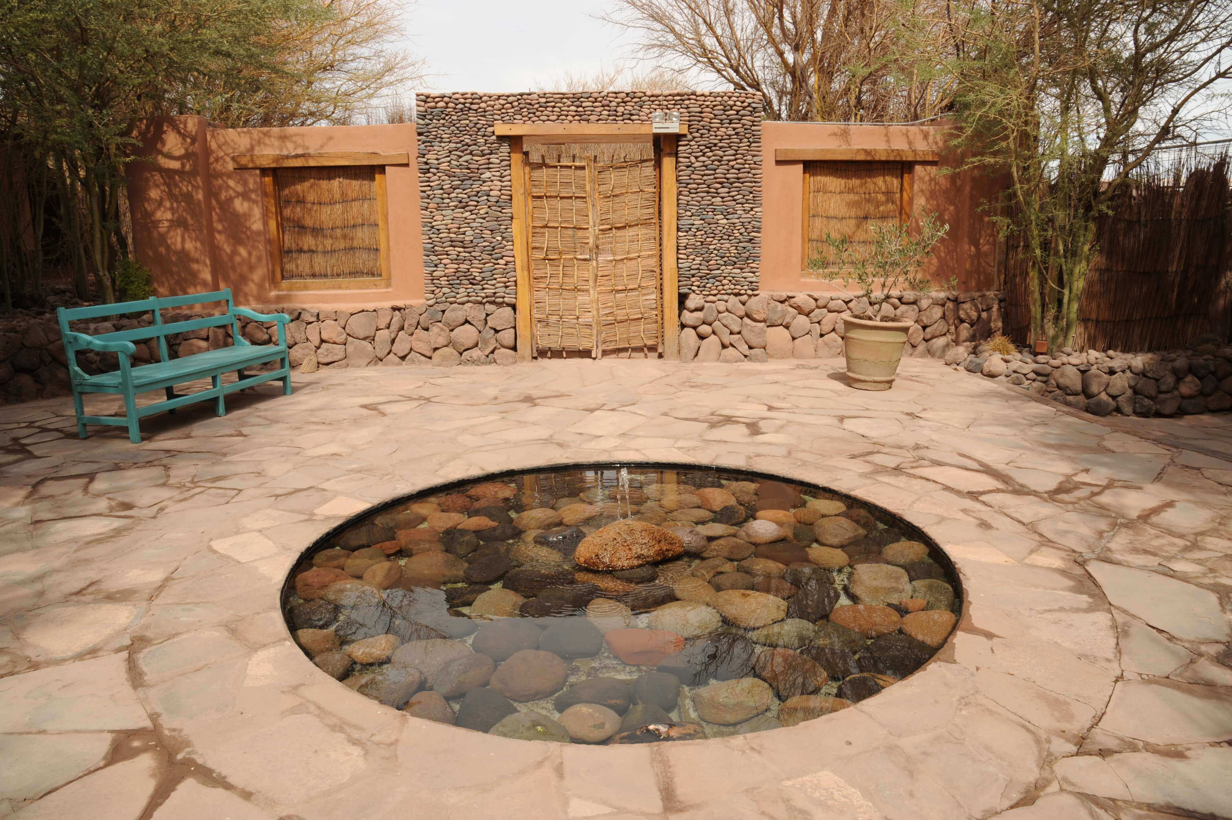 A peaceful stone courtyard in Chile's Atacama Desert features a circular pond, rustic wooden doors, and a turquoise bench. 