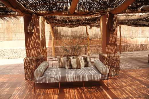 A cozy lounge with rustic cushions sits beneath a woven shade roof in the warm desert ambiance of Chile's Atacama region. 