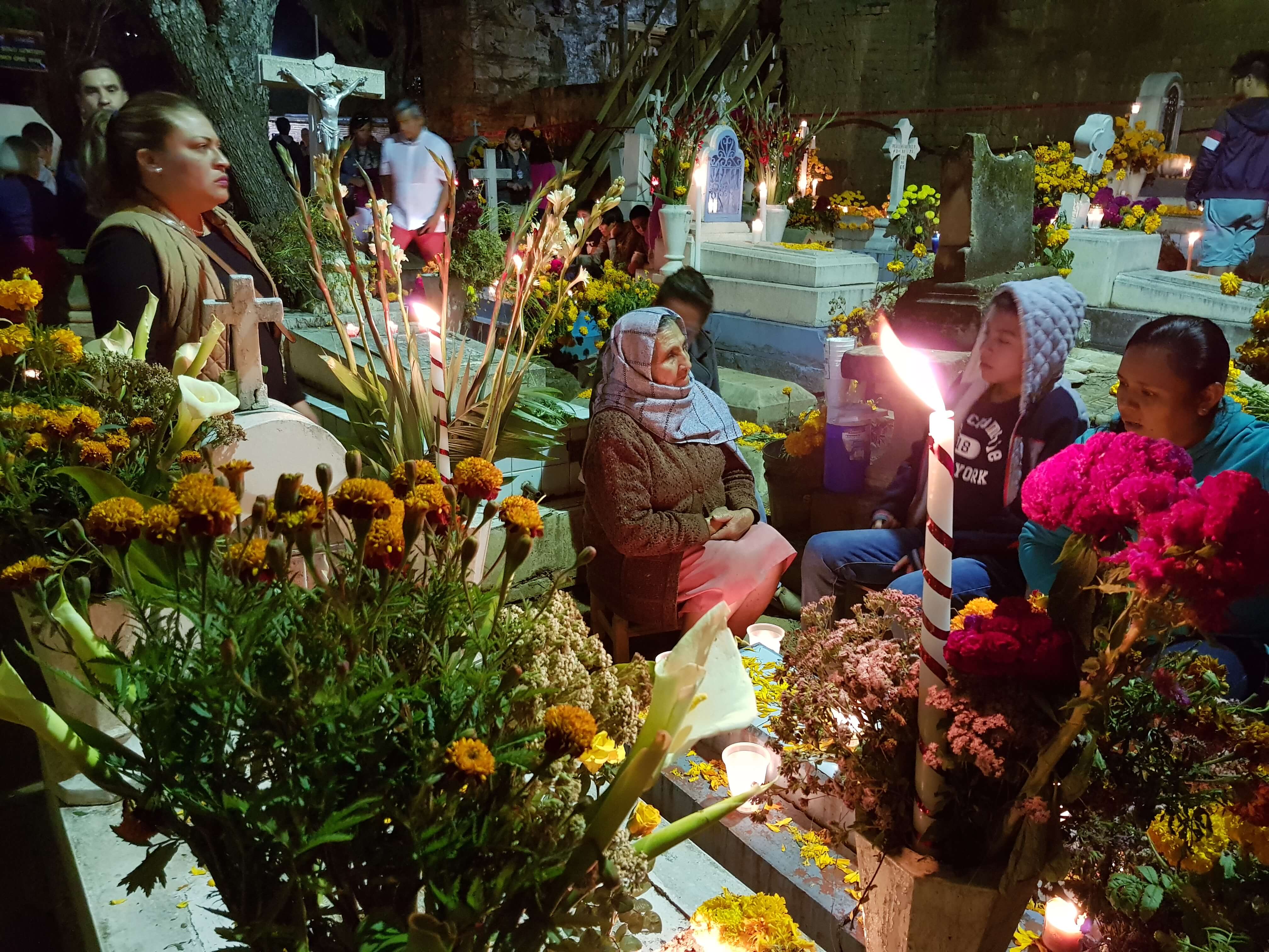 Families holding a night vigil at a cemetery in Mexico for Day of the Dead, surrounded by marigolds, candles, and offerings. 