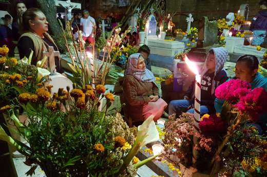 Families holding a night vigil at a cemetery in Mexico for Day of the Dead, surrounded by marigolds, candles, and offerings. 