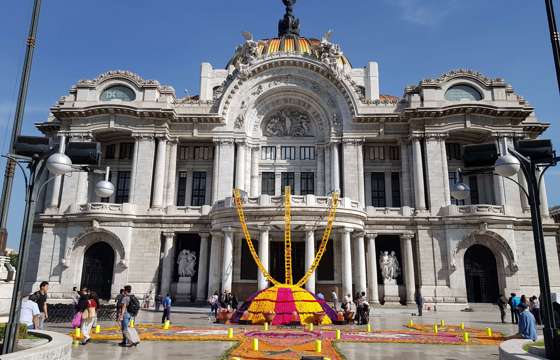 Palacio de Bellas Artes in Mexico City decorated with vibrant Day of the Dead marigold flowers and altars, under a clear blue sky. 