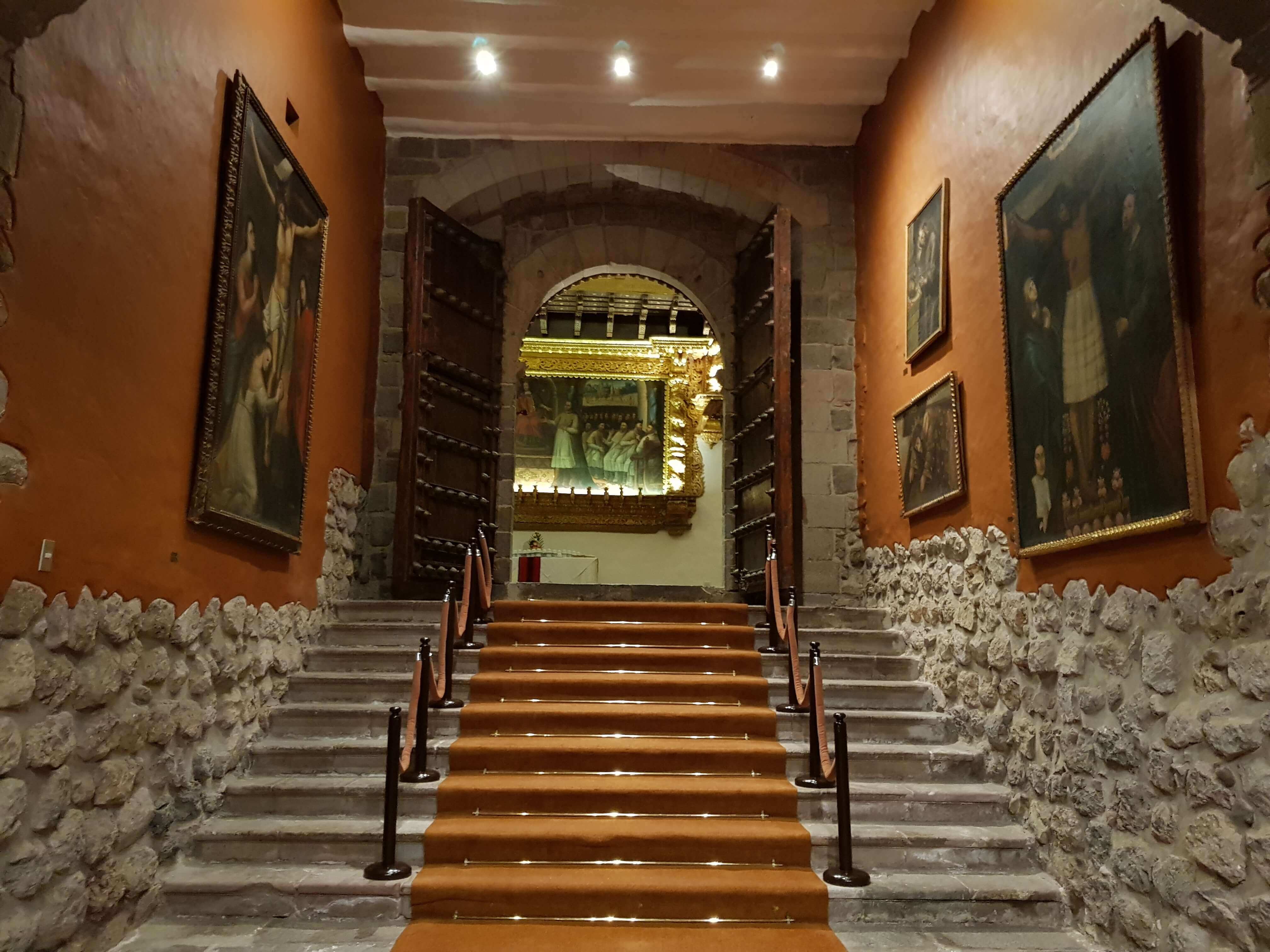 Interior of a colonial-era convent in Cusco, Peru, with stone walls, artwork, and wooden doors. 