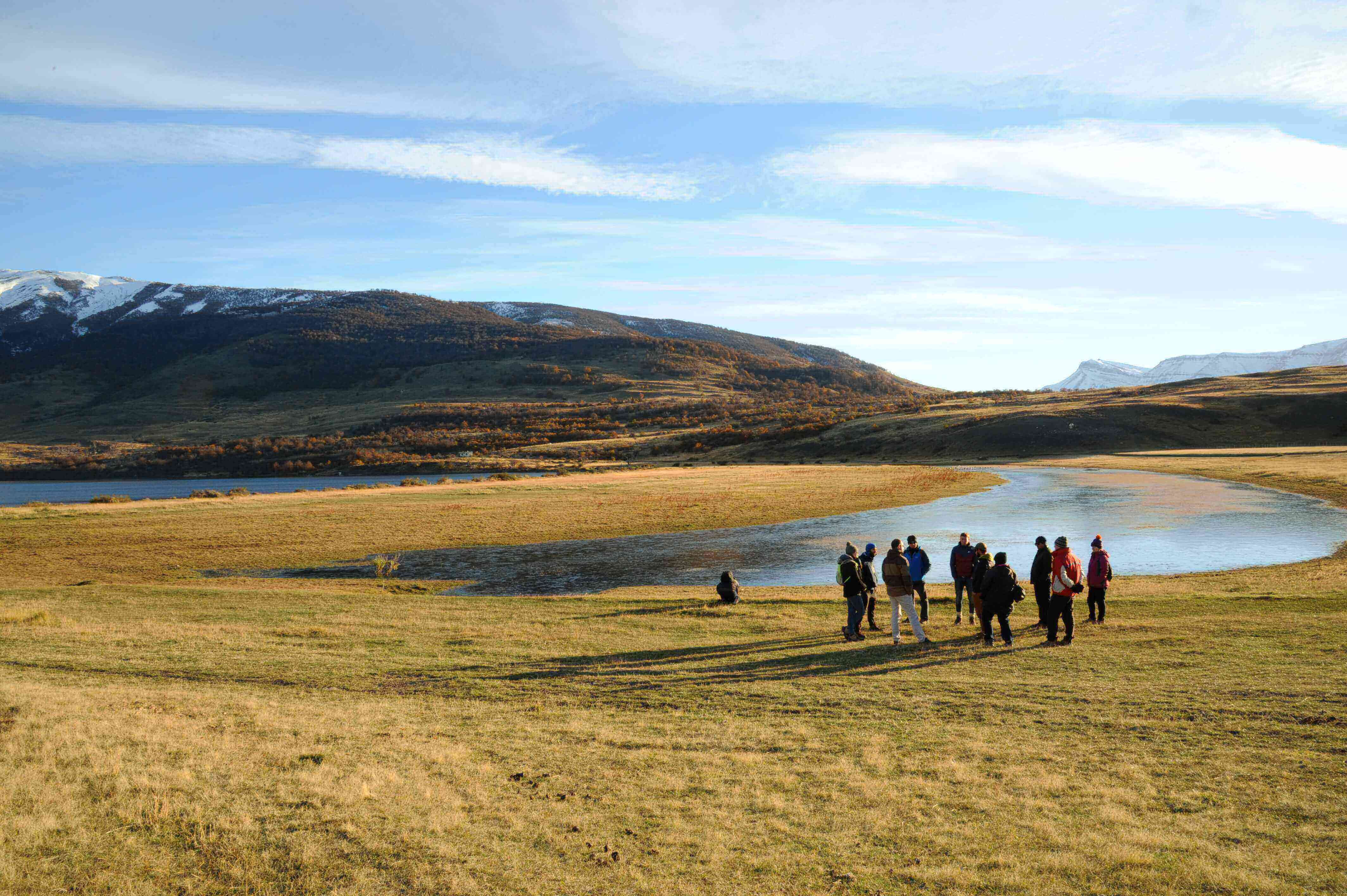 Travelers hiking across Patagonian grasslands near a lagoon in Torres del Paine, Chile, under a wide blue sky. 