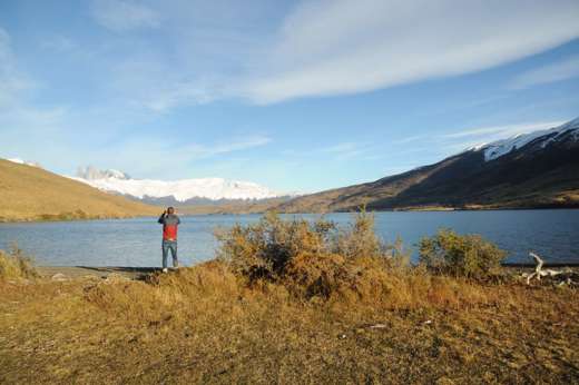 A traveler photograph the serene lake and jagged snow-capped peaks of Torres del Paine in Chilean Patagonia under a wide blue sky. 