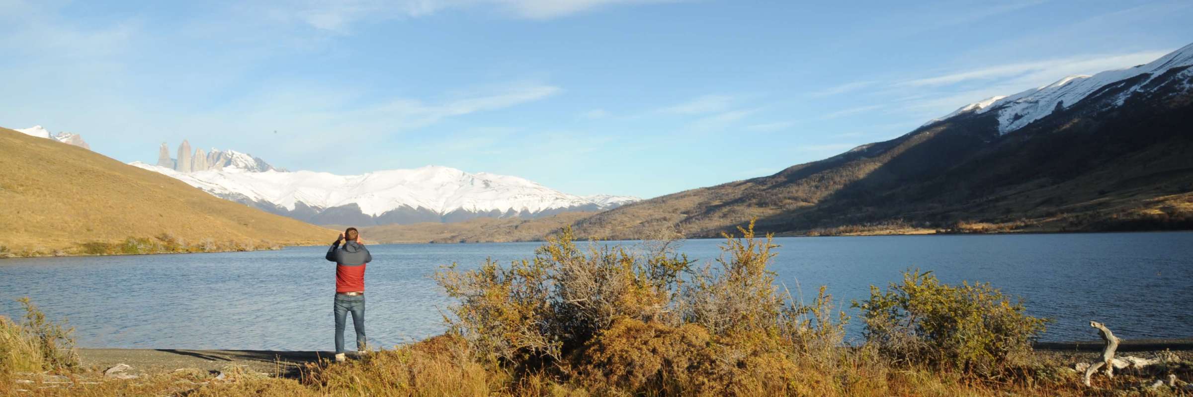 A traveler photograph the serene lake and jagged snow-capped peaks of Torres del Paine in Chilean Patagonia under a wide blue sky. 