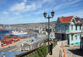 A turquoise colonial house overlooks Valparaiso's harbor, with cargo ships, hills, and vivid port activity beneath blue skies. 