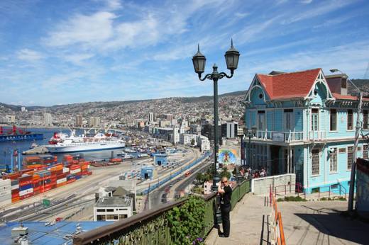 A turquoise colonial house overlooks Valparaiso's harbor, with cargo ships, hills, and vivid port activity beneath blue skies. 