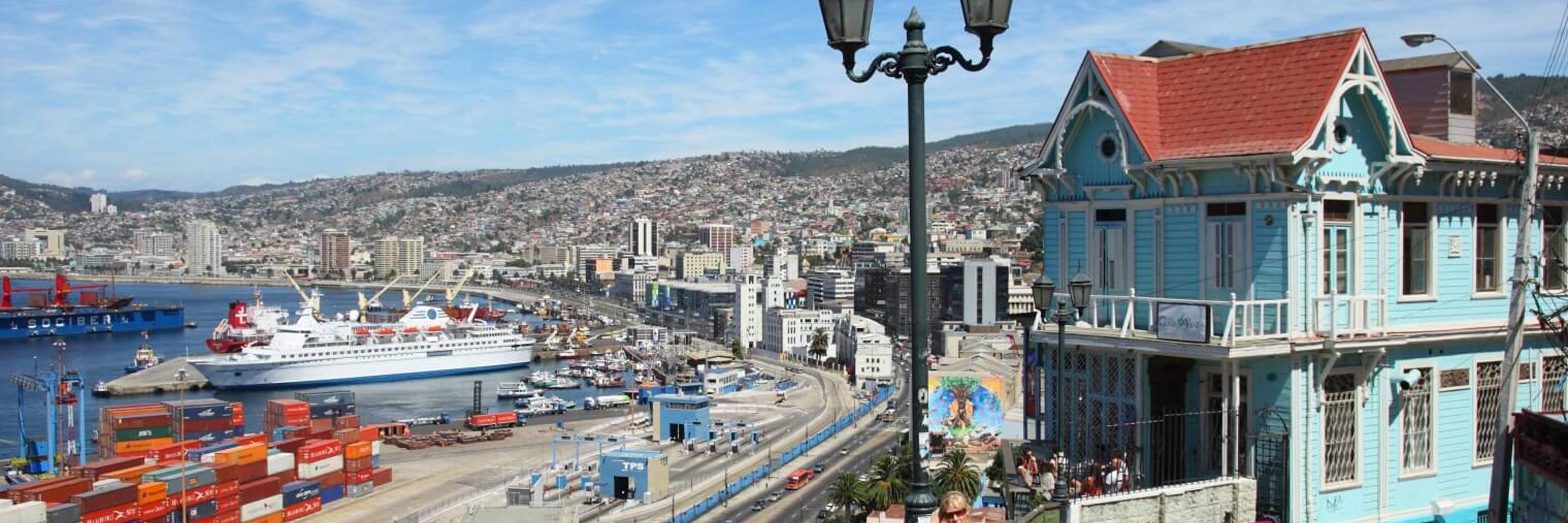 A turquoise colonial house overlooks Valparaiso's harbor, with cargo ships, hills, and vivid port activity beneath blue skies. 