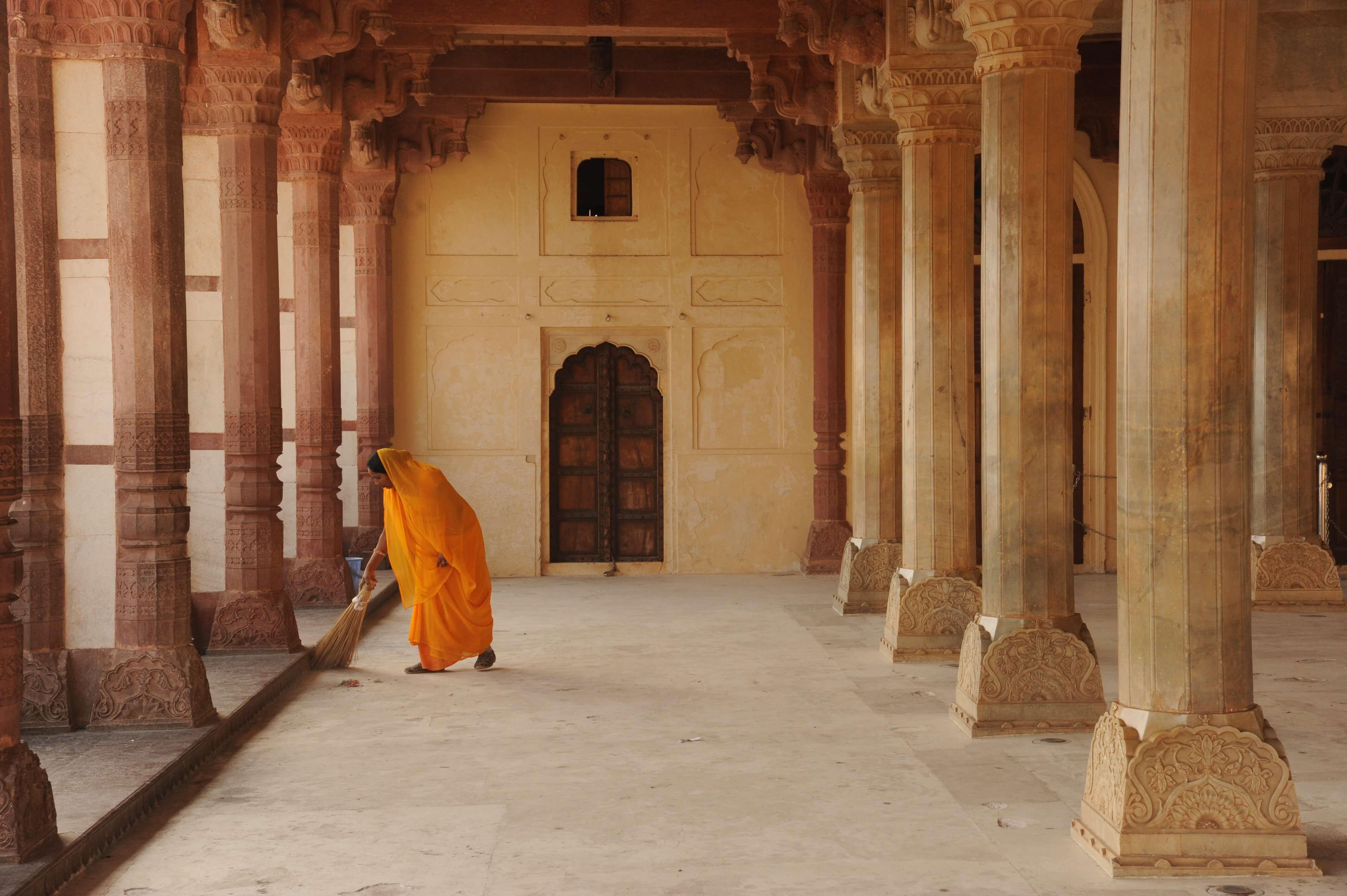 India- A person in a yellow robe sweeps the corridor of an ornately carved historic palace. 