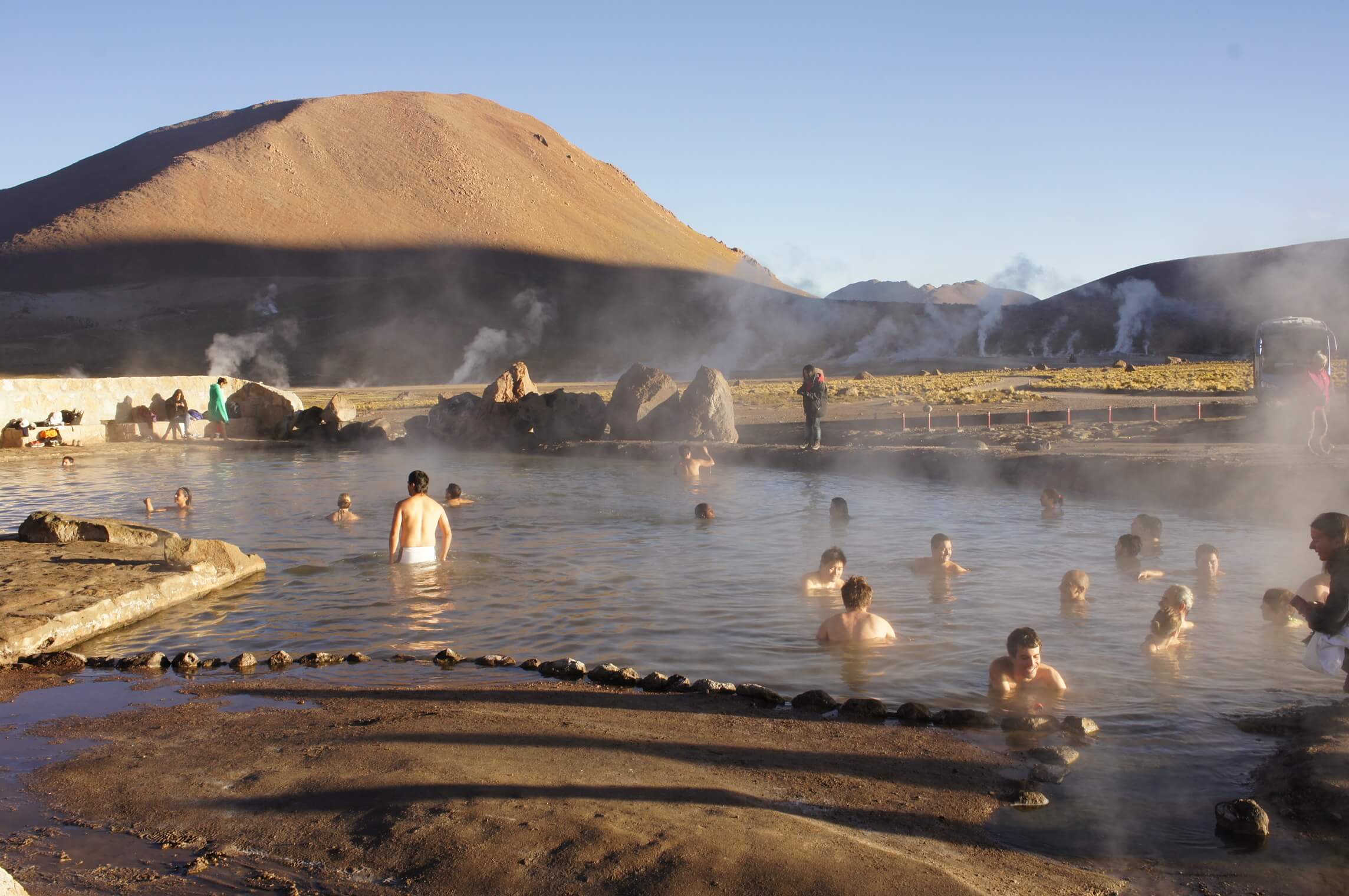 Travelers soak in El Tatio hot springs, surrounded by Andean peaks and rising steam near San Pedro de Atacama. 