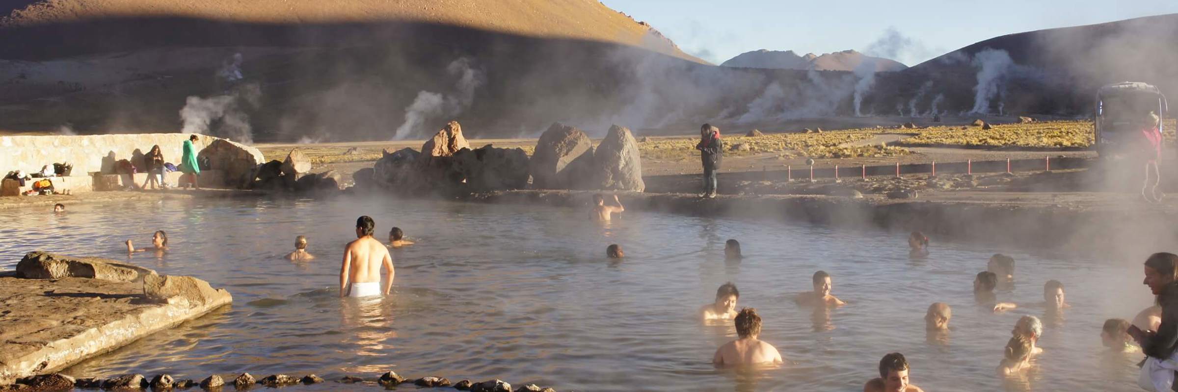 Travelers soak in El Tatio hot springs, surrounded by Andean peaks and rising steam near San Pedro de Atacama. 